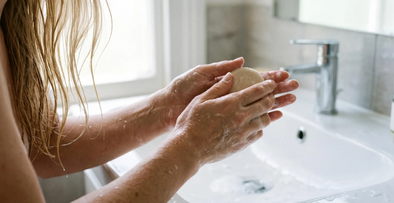 Des mains moussent un shampoing solide au-dessus d'un lavabo en céramique blanche.